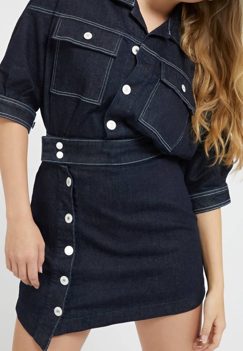 Robe en denim bleu foncé avec des boutons blancs, des poches avant et une ceinture à la taille, portée par une femme aux longs cheveux ondulés.