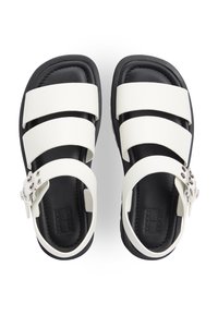 Λευκές σανδάλια με λουράκια και μαύρη σόλα, με φαρδιές λωρίδες και ασημένια αγκράφα. Ομαλό υλικό με αντίθετη υφή.