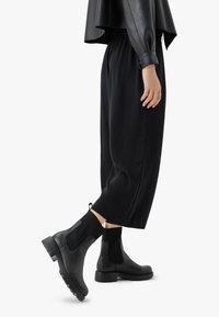 Črne čevlje Chelsea z elastičnimi stranskimi paneli, teksturiranimi pletenimi detajli in gumijastimi podplati, v kombinaciji z ohlapnimi črnimi hlačami in črnim vrhom.