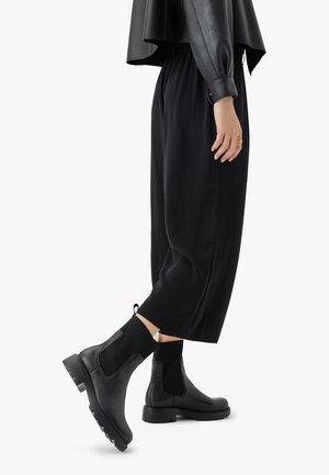 Black chelsea boots with elastic side panels, textured knit accents, and rubber soles, paired with loose black trousers and a black top.