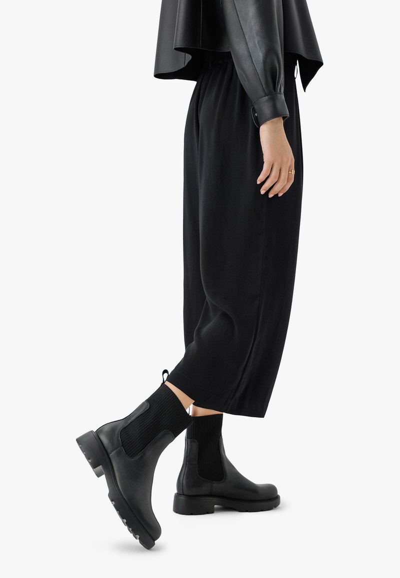 Črne čevlje Chelsea z elastičnimi stranskimi paneli, teksturiranimi pletenimi detajli in gumijastimi podplati, v kombinaciji z ohlapnimi črnimi hlačami in črnim vrhom.