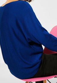 Maglietta blu, a maniche lunghe e dal taglio ampio con scollo largo, abbinata a pantaloni neri, seduta su una sedia rosa. Tessuto liscio, design casual.