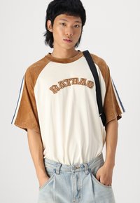 T-shirt oversize bicolore blanc et marron avec le texte "RATBAG". Doté de manches courtes avec des rayures bleues, en tissu doux et coupe décontractée.