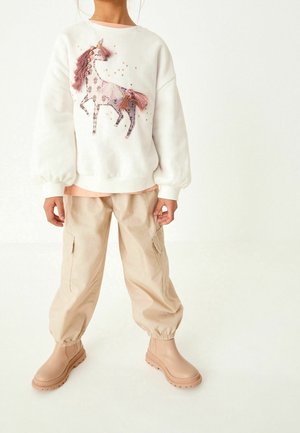 Enfant portant un sweat-shirt blanc avec un cheval rose décoratif, un pantalon cargo beige et des bottes beige, debout devant un fond uni.