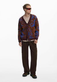 Cárdigan de punto en marrón con patrones de paisley azul, con escote en V, cierre de botones y puños acanalados. Combinado con pantalones cargo oscuros y zapatillas.