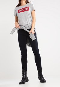 Mujer con una camiseta gris de Levi's, jeans skinny negros, botas de tobillo negras con cordones y un suéter gris atado a la cintura.