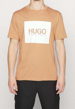 Homme portant un T-shirt beige à manches courtes avec un carré blanc et le texte "HUGO HUGO BOSS" à l'avant, associé à un pantalon noir.