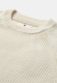 Strikket sweater i cremefarve med ribbet struktur, rund halsudskæring og blød finish. Har en synlig søm langs skuldrene og kraven.