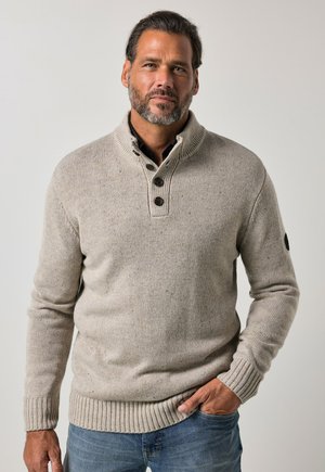 Homme aux cheveux foncés et à la barbe portant un pull beige en maille avec des boutons et un jean bleu, debout avec une main dans la poche.