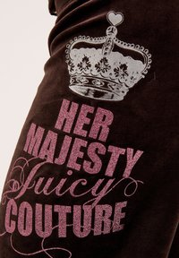 Brauner Stoff mit einer strukturierten Oberfläche, der ein silbernes Kronenmotiv zeigt. Glitzernder pinker Text lautet "IHRE MAJESTÄT Juicy Couture."