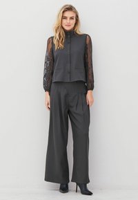 Woman wearing black buttoned top with lace sleeves, wide-leg black trousers, and black heeled ankle boots, standing against a white wall.