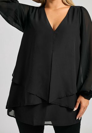 Woman wearing a black layered long-sleeve blouse with a V-neck, standing with one hand raised and the other resting by her side.