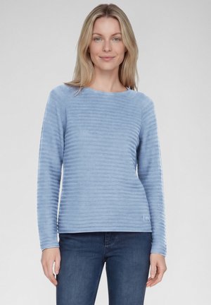 Woman with blonde hair wearing a light blue ribbed long-sleeve top and dark blue jeans, standing against a plain background.