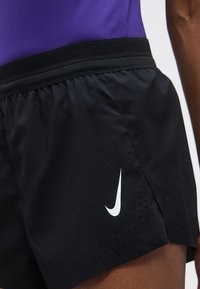 Κοντινή λήψη μαύρων αθλητικών σορτς με διάτρητα σημεία και λευκό λογότυπο Nike Swoosh στο πλάι, που φοριούνται με μοβ τοπ.