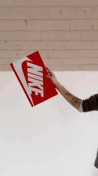 Červená krabice na boty Nike s bílým logem, obdélníkový tvar, lesklý povrch, držená v jedné ruce na jednobarevném bílém pozadí.