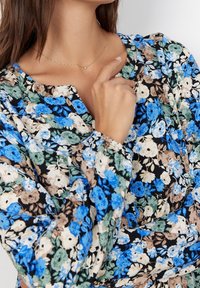 Blommig blus med svart bakgrund som har blå, gröna och beige blommor, smockade ärmar och rund halsringning. Ljusgul halsband.