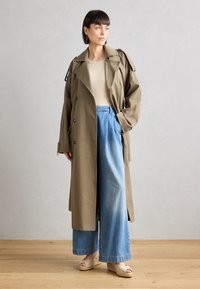 Larga trench verde oliva con un diseño de doble botonadura, acompañada de anchos pantalones de mezclilla azules. El conjunto presenta sandalias de un ligero color beige.