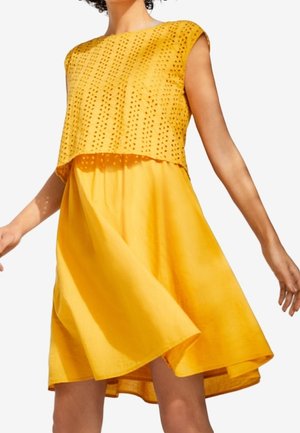 Kobieta w żółtej sukience bez rękawów z perforowaną teksturowaną nakładką na górze, w ruchu, powodująca rozchylanie się spódnicy na zewnątrz.
