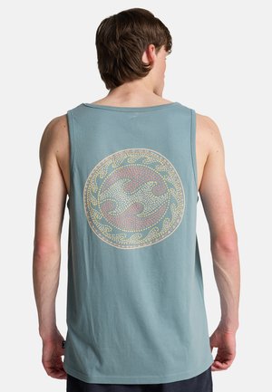 Jonge man die een lichtblauw mouwloos shirt draagt met een cirkelvormig golfpatroon op de rug, staand tegen een effen achtergrond.