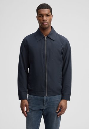 Giovane uomo con capelli corti e ricci che indossa una giacca blu scuro con cerniera e jeans blu, in piedi davanti a uno sfondo semplice grigio chiaro.