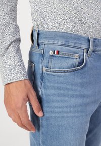 Világoskék farmernadrág ötpocketes dizájnnal, réz szemcsékkel, egy kis zsebbel piros, fehér és kék részletekkel, valamint derékpánttal.