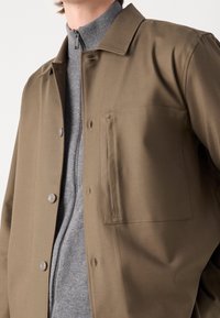 Chaqueta verde oliva con un acabado suave, cierre de botones y un bolsillo en el pecho. Lleva puesta sobre un suéter gris con cremallera de textura acanalada.