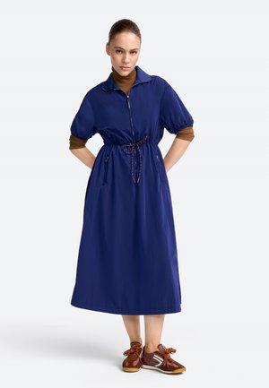 Kobieta ubrana w granatową sukienkę midi z suwakiem i talią ze sznurkiem, pod spodem brązową koszulę z długim rękawem oraz brązowe trampki ze sznurówkami w kolorze pomarańczowym.