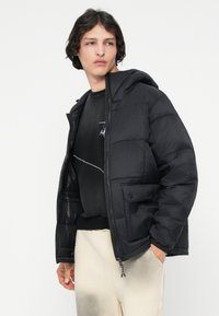 Μαύρο μπουφάν με κουκούλα, με κεντημένο σχέδιο και μπροστινό φερμουάρ. Φοριέται πάνω από ένα σκούρο φούτερ και κρεμ παντελόνι.