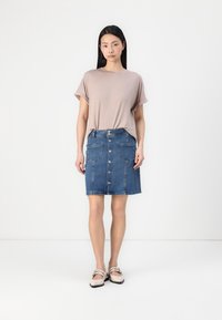 Kortermet beige t-skjorte kombinert med et blått denimskjørt med fremre lommer og sølvknapper nedover midten. Jevn stofftekstur.