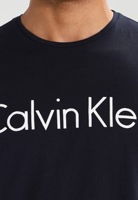 T-shirt en coton navy arborant le texte "Calvin Klein" en grandes lettres blanches, col rond et manches courtes. Texture lisse.