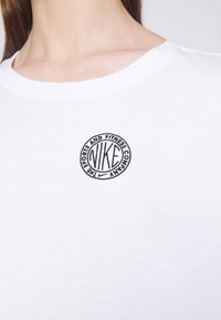 Vit bomull T-shirt med en rund svart broderad logotyp med texten "NIKE" och "THE SPORTS AND FITNESS COMPANY" runt om. Enkel rund halsringning.