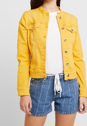 Žena v žluté bundě na knoflíky přes bílou košili a modré džínové šortky s modrými proužky, stažené bílým pruhem s pruhy.