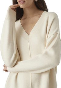 Pull en tricot beige avec un décolleté en V profond, des manches larges et des poignets côtelés ; texture douce sans motifs ni matériel visible.
