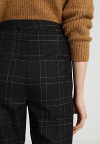 Černé kalhoty s kostkovaným vzorem a texturovanou tkaninou, s hnědo-šedým károvaným vzorem. Pas je na míru a vzadu je kapsa.