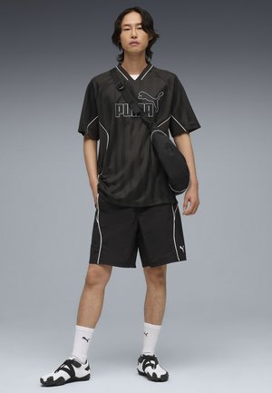 Młody mężczyzna stoi, ubrany w czarną odzież sportową Puma, białe skarpetki Puma, czarno-białe buty oraz czarną torbę na ramię na szarym tle.