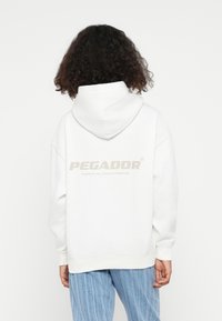 Weißer Kapuzenpullover mit großem hellbeigem "PEGADOR"-Logo auf dem Rücken. Der Stoff sieht weich aus und hat eine lockere Passform sowie gerippte Bündchen.