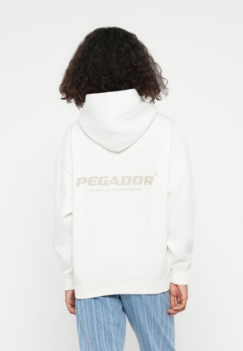 Weißer Kapuzenpullover mit großem hellbeigem "PEGADOR"-Logo auf dem Rücken. Der Stoff sieht weich aus und hat eine lockere Passform sowie gerippte Bündchen.