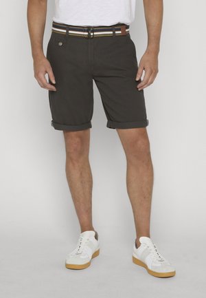 Mann trägt dunkle hochgekrempelte Shorts, weiße Sneakers mit Gummisohlen und einen gestreiften Gürtel, steht vor einem einfarbigen Hintergrund.