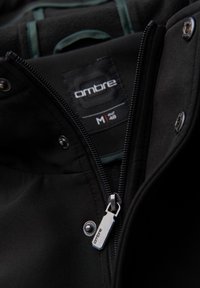Chaqueta negra con cremallera y cierre de botón a presión. La etiqueta dice "ombre". Cuenta con un tejido suave y un forro interior en contraste.