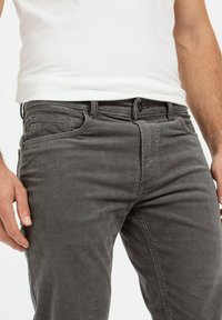 Grijze corduroy broek met een gestructureerd oppervlak, vijf zakken en een knoopsluiting bij de tailleband. Eén hand aan de zijkant geplaatst.