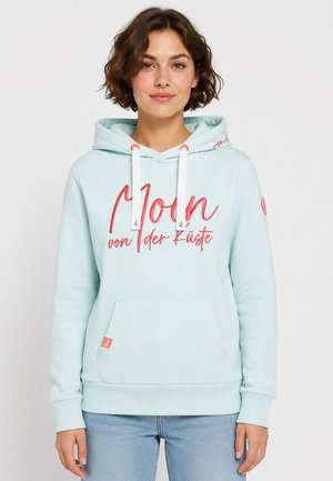 Jonge vrouw met kort bruin haar die een lichtblauwe hoodie met witte trekkoorden en rode tekst "Moon von der Küste" en lichtblauwe spijkerbroek draagt.
