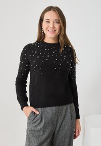 Czarny sweter dzianinowy z perełkowymi zdobieniami na przodzie, prążkowanymi mankietami i dołem, stylizowany z szarymi, plisowanymi spodniami.