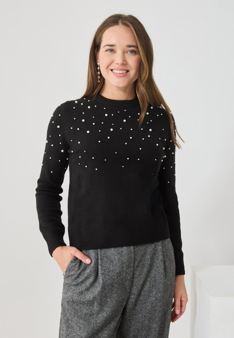 Czarny sweter dzianinowy z perełkowymi zdobieniami na przodzie, prążkowanymi mankietami i dołem, stylizowany z szarymi, plisowanymi spodniami.