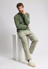 Zielony sweter z dzianiny z guzikami na ramionach, połączony z jasnobeżowymi spodniami typu cargo i kremowymi sneakersami, siedzący na białym piedestale.