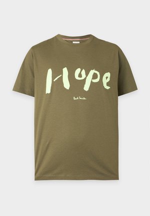 Homme debout les mains dans les poches, portant un T-shirt vert olive avec le texte « Hope », un short ample foncé, des chaussettes blanches et des baskets blanches.