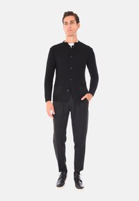 Cardigan nero a costine con scollatura rotonda, sei bottoni e due tasche frontali, abbinato a pantaloni neri e scarpe nere.