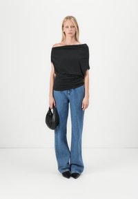 Zwarte off-the-shoulder top met gedrapeerd ontwerp, gecombineerd met lichtblauwe wijde denim jeans. Het model houdt een kleine zwarte tas vast en draagt puntige schoenen.
