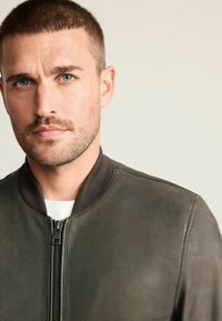 Tamnozeleni kožni bomber jakna s rebrastim ovratnikom i zatvaranjem na patentni zatvarač. Glatka tekstura, minimalni detalji i prilagođeni dizajn.