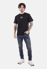 T-shirt nera in cotone con stampa grafica sul petto, abbinata a jeans di denim blu e sneakers grigie. Uomo in una posa neutra.