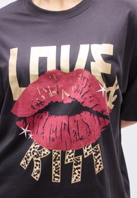 Nærbilde av en svart T-skjorte med stor rød leppegrafikk over gullfarget "LOVE"-tekst og leopardmønstret "KISS"-bokstaver under.